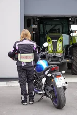 Fahrschülerin steht neben einem Motorrad und trägt eine Fahrschulbekleidung mit dem Hinweis „Fahrschule“.